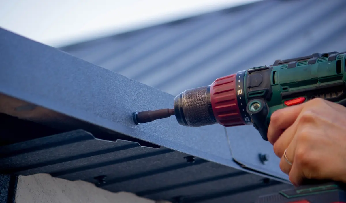 A hand uses a cordless drill on a dark metal roofing material. The drill is red and green, and the screw is being driven into the metal.