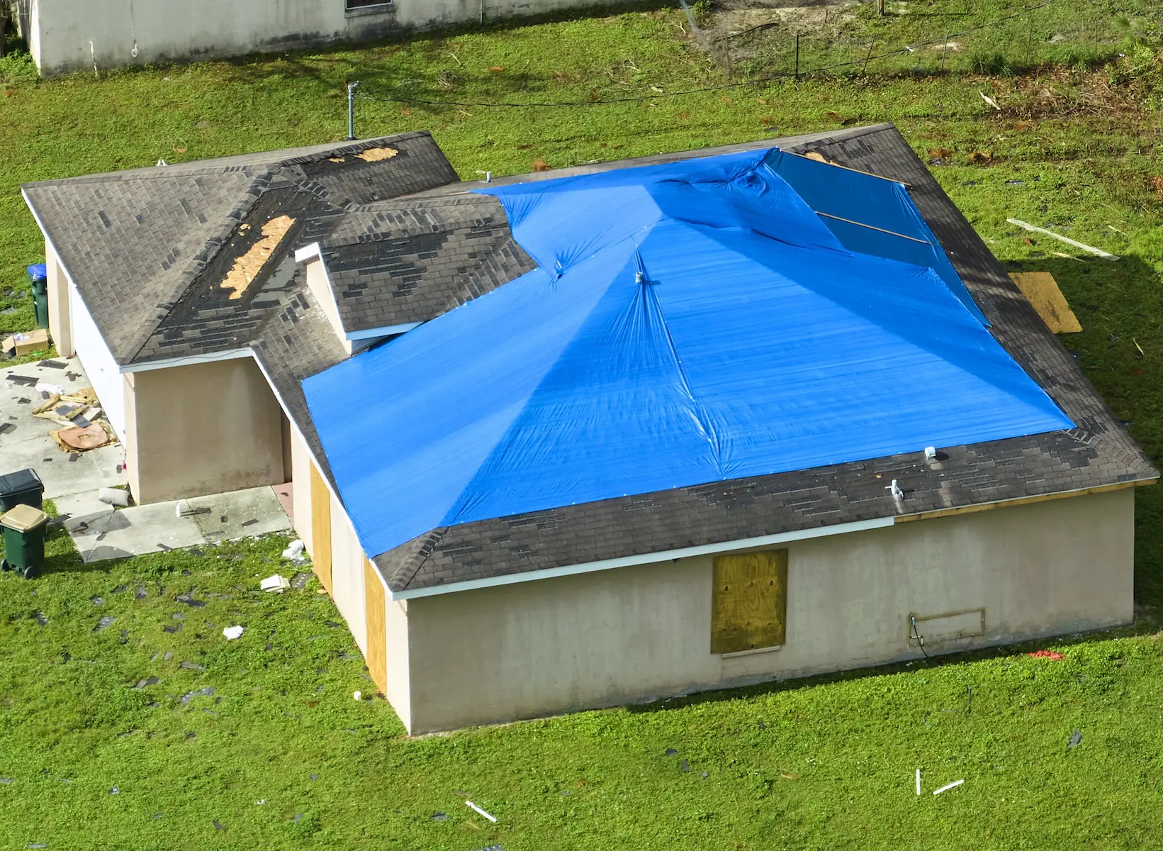 An aerial shot captures a house in a suburban setting. A blue tarp covers a portion of the roof that has damaged or missing roof tiles. The yard is green. Some windows are boarded up with plywood.